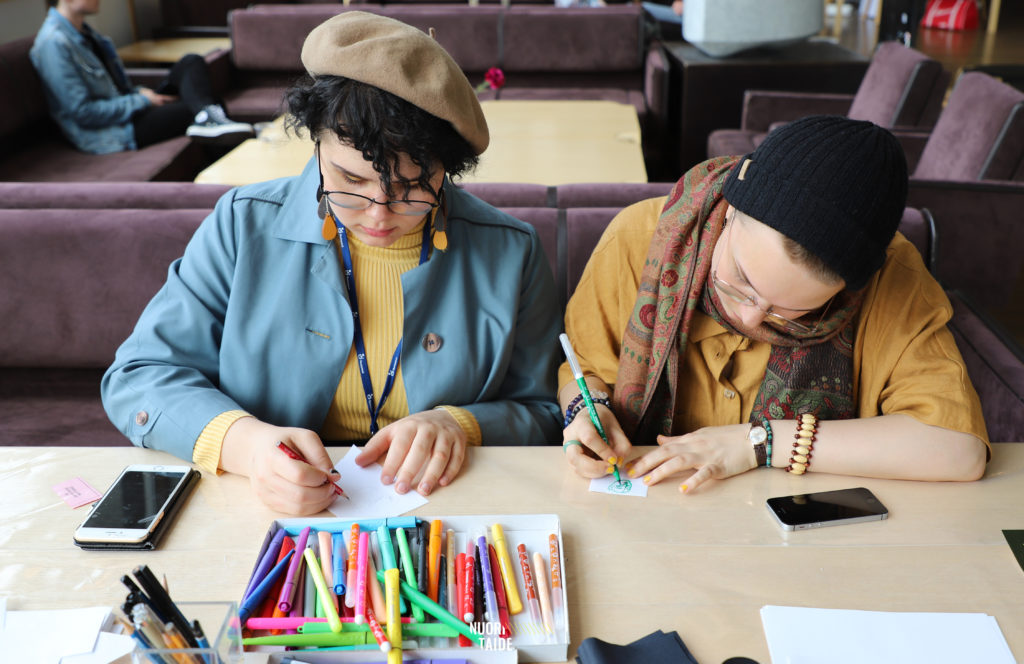 Kaksi nuorta piirtää pienille paperilapuille. Pöydällä on kyniä, papereita ja kaksi puhelinta. Taustalla sohvaryhmä jossa istuu yksi henkilö.