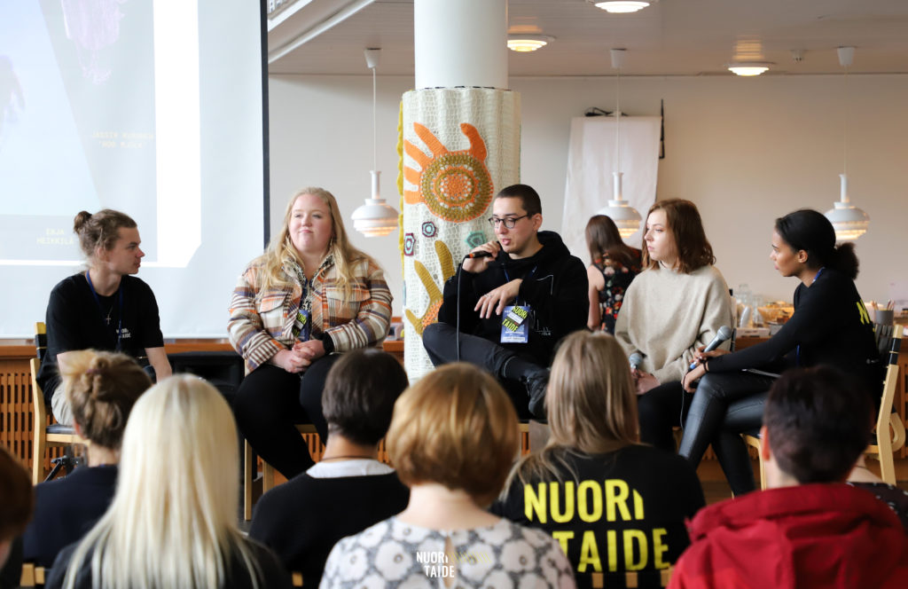 Selkäpuolelta kuvattuja henkilöitä, yhdellä henkilöllä t-paita jonka selkäpuolella Nuori Taide-logo. He katsovat edessä olevaa keskusteluryhmää.