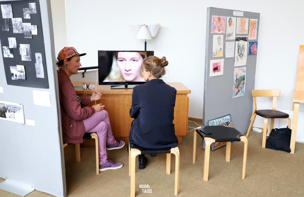 Kaksi henkilöä istumassa jakkaroilla. Katselevat tv-ruutua jossa on kuva henkilön kasvoista.