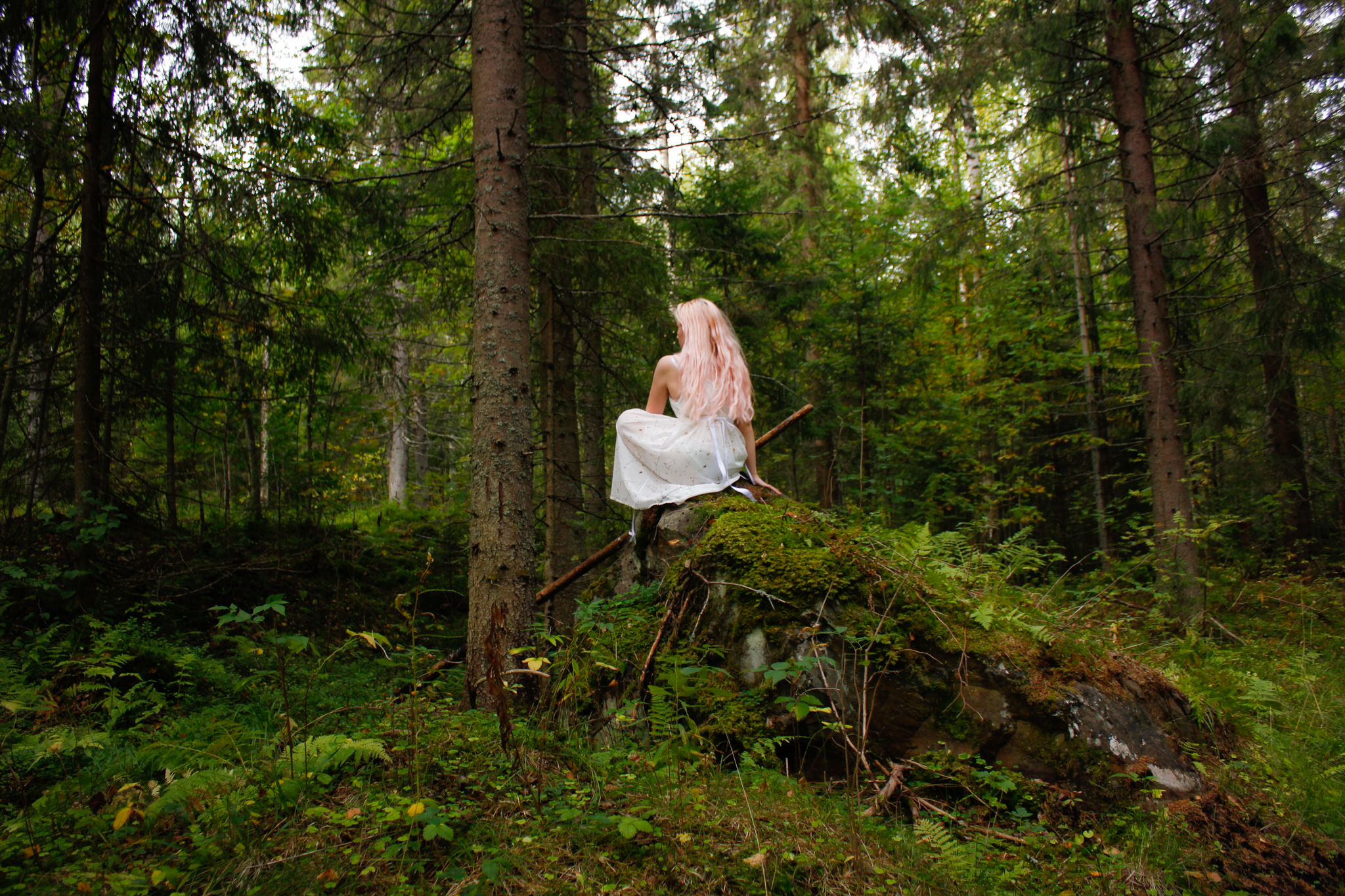 Henkilö istuu kiven päällä metsässä selkä kuvaajan päin. Hänellä on yllään mekko ja pitkät hiukset auki selkää vasten.