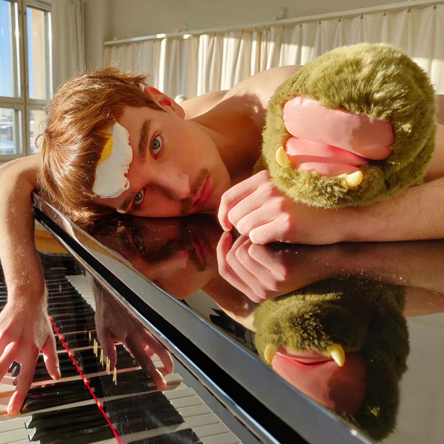 Henkilö makaa pianon päällä ja hänellä on otsan päällä kananmuna ja kainalossa pehmo.