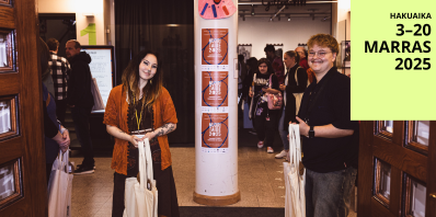 Kaksi henkilöä seisoo sisääntuloaulassa. Taustalla taidetapahtuman kävijöitä