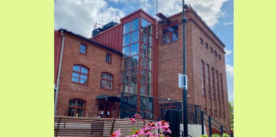 Kuva tiilirakennuksesta poutapäivänä. Kuvan seunoilla yksiväriset palkit rajaamassa kuvaa