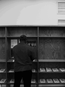 Henkilö seisoo selin kuvaajaan hyllyn edessä, joissa hyllylevyillä pieniä kirjoja pinoissa